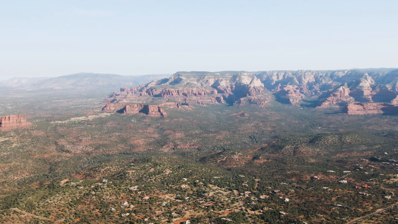 High aerial frame of Sedona, Arizona showing a scrub-covered valley, small roads and buildings below, red rock cliffs across the background, and no visible people.