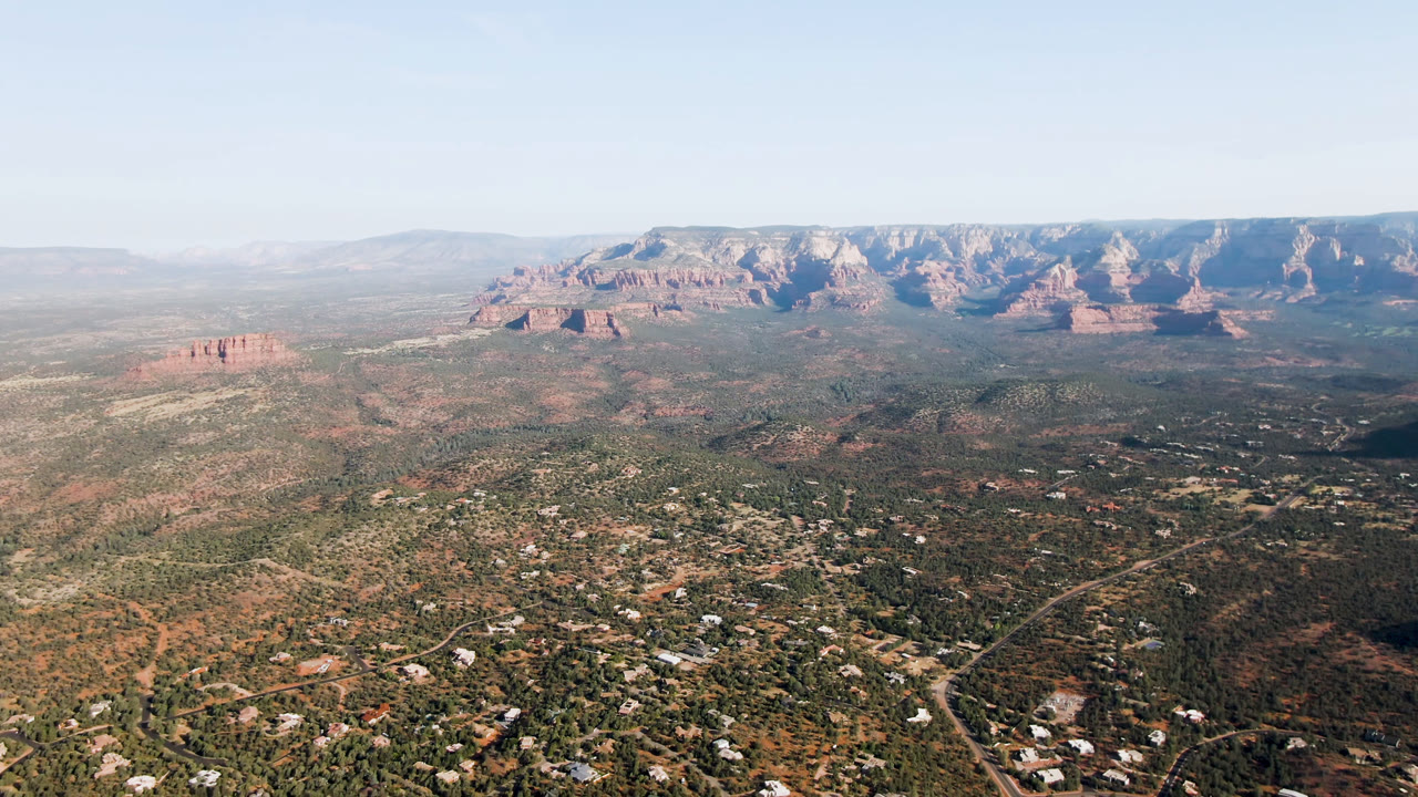 Second frame of the Sedona aerial view, with residential roads and scrub terrain below, a small red rock formation to the left, distant cliffs, and no visible people.
