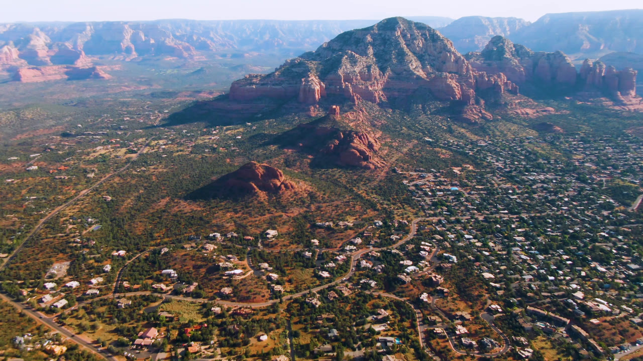 Aerial frame over Sedona, Arizona, showing denser city blocks in the lower frame, a rounded red butte near center, larger cliffs behind it, and no visible people.