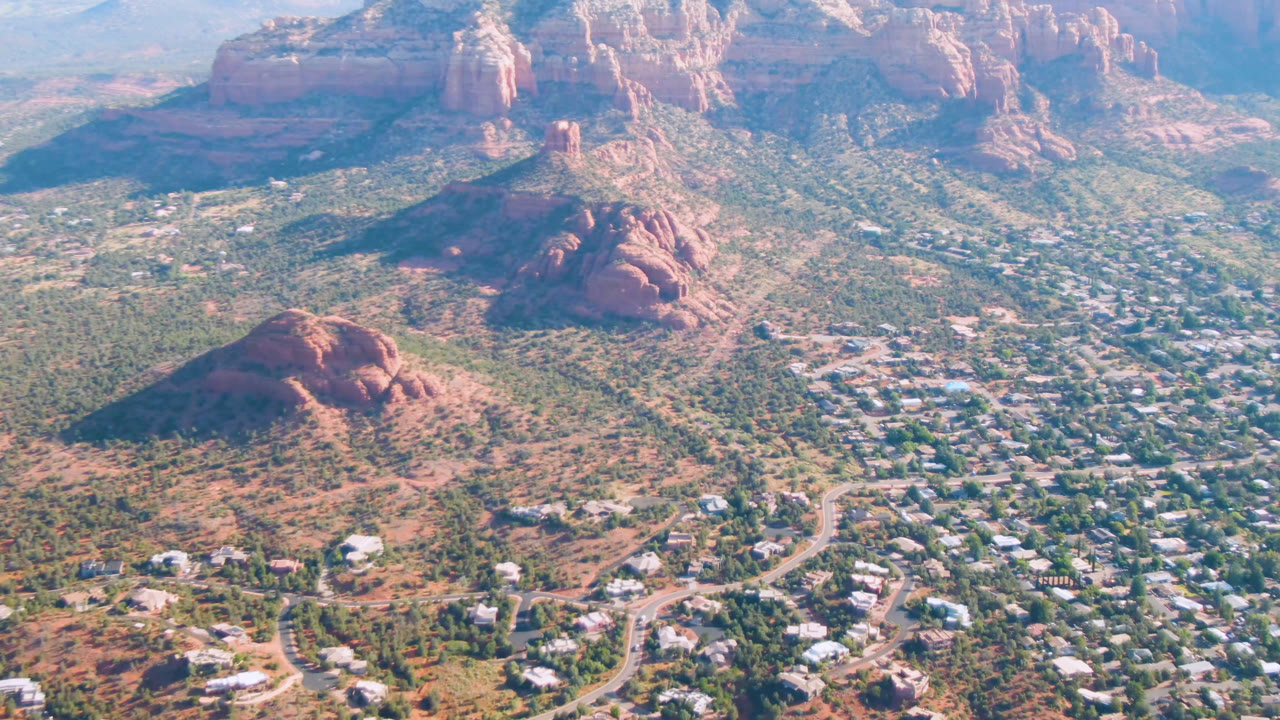 Aerial frame of Sedona, Arizona showing a rounded red rock formation near the center, residential roads and buildings below, hazy cliffs beyond, and no visible people.