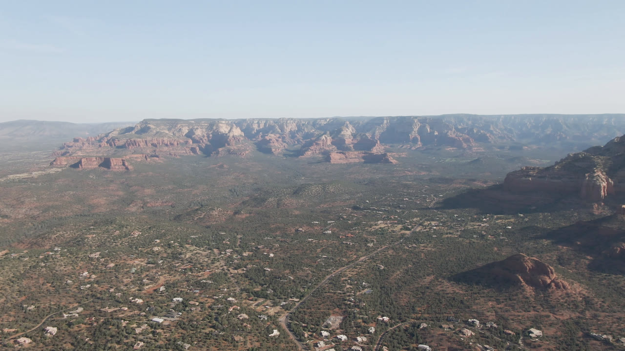 A high aerial view of Sedona, Arizona, with scrub-covered valley terrain, small buildings, branching roads, red rock formations, distant ridges, clear sky, and no visible people.
