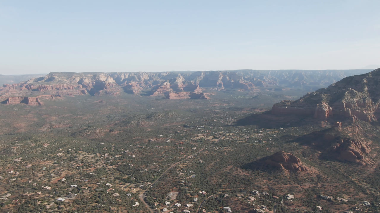 Opening frame of Sedona, Arizona from above, showing a broad desert valley, scattered buildings and roads, distant red rock formations, a right-side mountain, and no visible people.