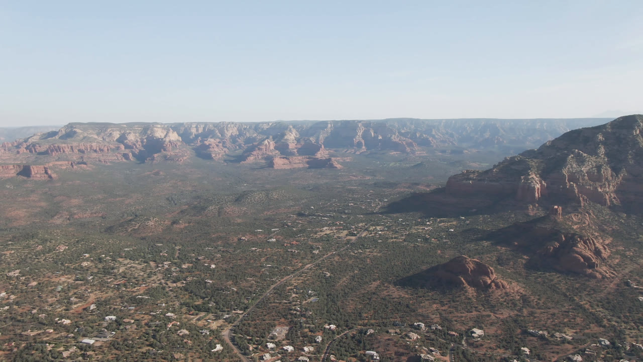 Second frame of the Sedona aerial view, with the dark mountain mass more central, red rock mesas along the far horizon, desert vegetation below, and no visible people.