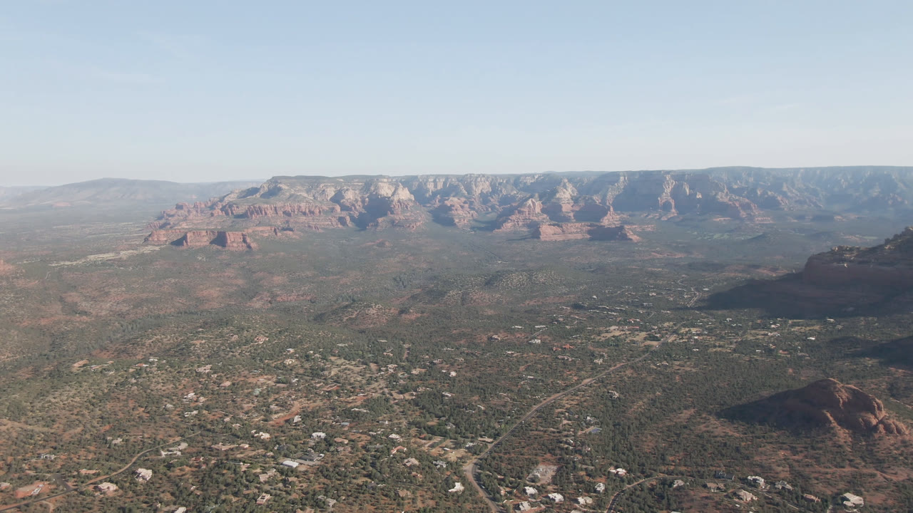 Opening frame of a Sedona, Arizona aerial view over a broad valley, scattered buildings, pale roads, red rock formations, distant mountain wall, and no visible people.