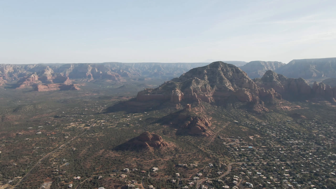 Second frame over Sedona, Arizona, with the central rock mass more prominent above the town grid, distant plateau walls, soft daylight, and no visible people.