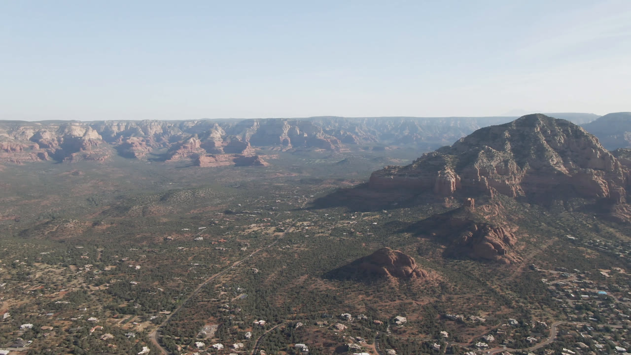 Opening aerial frame over Sedona, Arizona, showing a wide valley, scattered buildings, red rock formations, a central mountain mass, distant mesas, and no visible people.