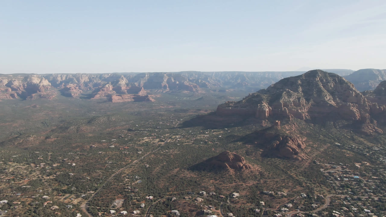 Second aerial frame in Sedona, Arizona, with the central mountain slightly left of center, red cliffs across the horizon, desert roads below, and no visible people.