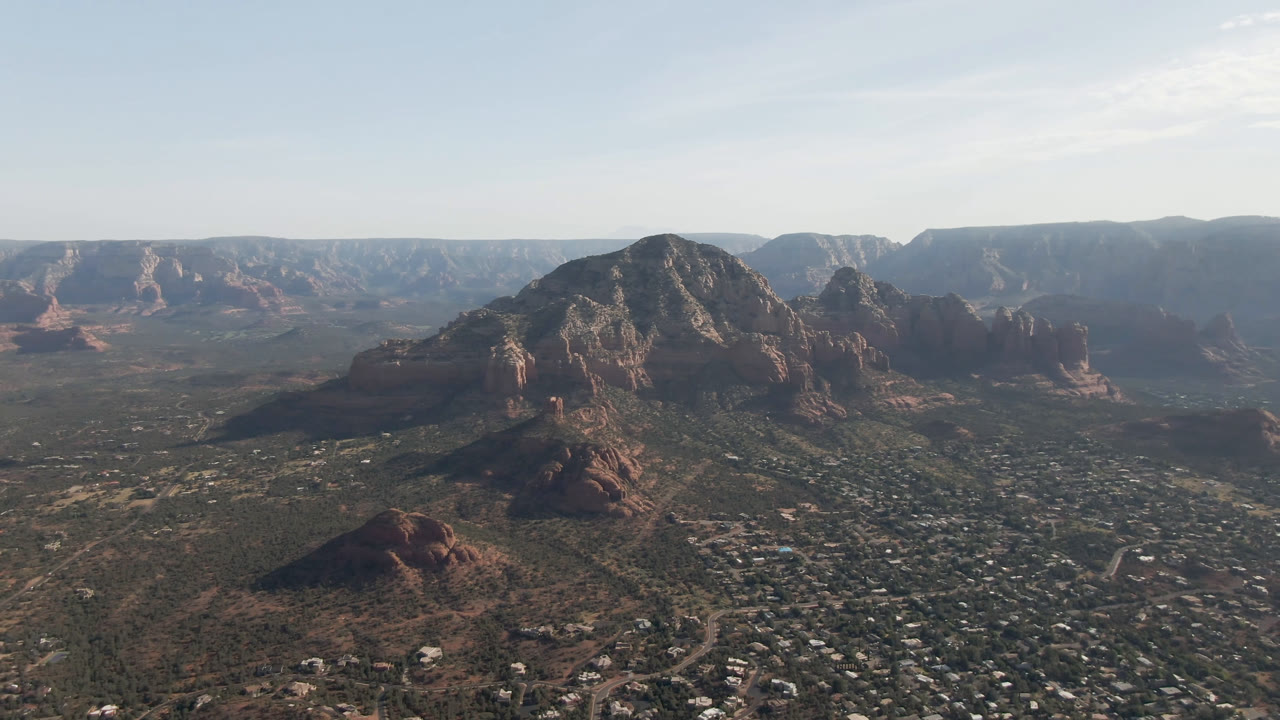 Opening aerial frame over Sedona, Arizona, with red rock buttes rising from a valley of scattered roads and buildings, no visible people.