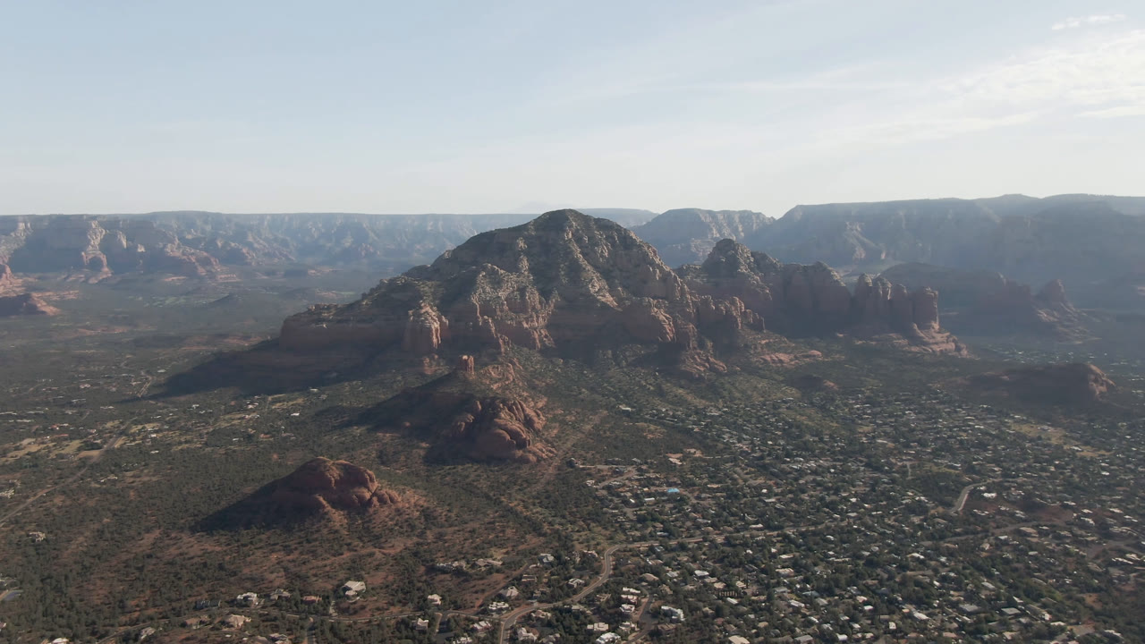 Sedona aerial view centered on a rounded red rock bluff, with valley neighborhoods in the lower frame and hazy mesas across the horizon, no visible people.