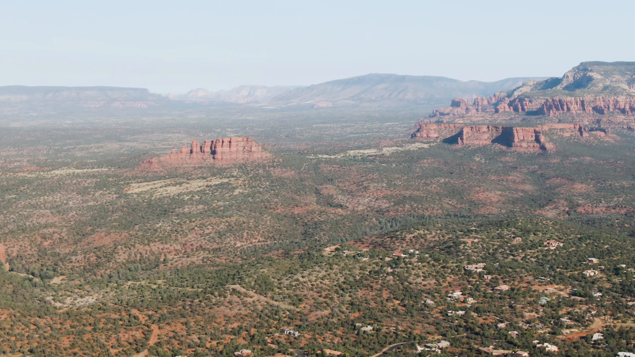 Wide aerial frame over Sedona, Arizona with scrub-covered valley, red rock bluffs, distant mesas, scattered low buildings, and no visible people.