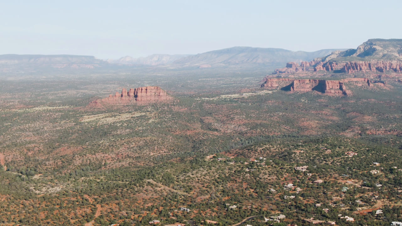 Sedona aerial view one second into the clip, showing a red butte left of center, a larger mountain formation in the background, broad scrubland, and no visible people.