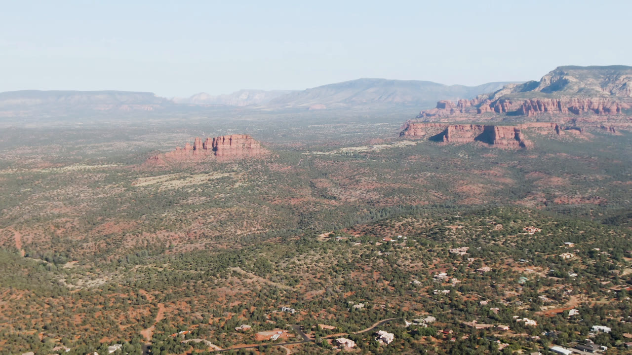 Opening aerial frame over Sedona, Arizona, showing scrubland, scattered homes, red rock buttes, distant ridges, pale blue sky, and no visible people.