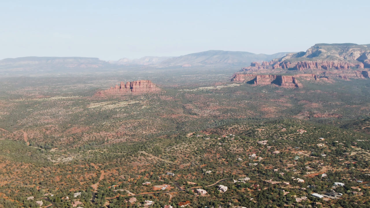 Second Sedona aerial frame with the small red rock formation shifted toward the lower left, broad desert terrain, hillside homes, distant cliffs, and no visible people.