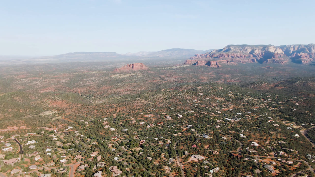 Wide aerial over Sedona, Arizona with dense desert vegetation, residential-looking structures, a small red rock butte left of center, and no visible people.