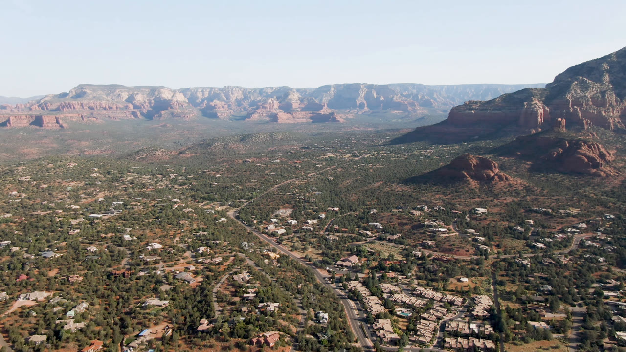Wide aerial view over Sedona, Arizona, showing roads, desert trees, scattered buildings, red rock formations, and a distant ridge under clear daylight, with no visible people.