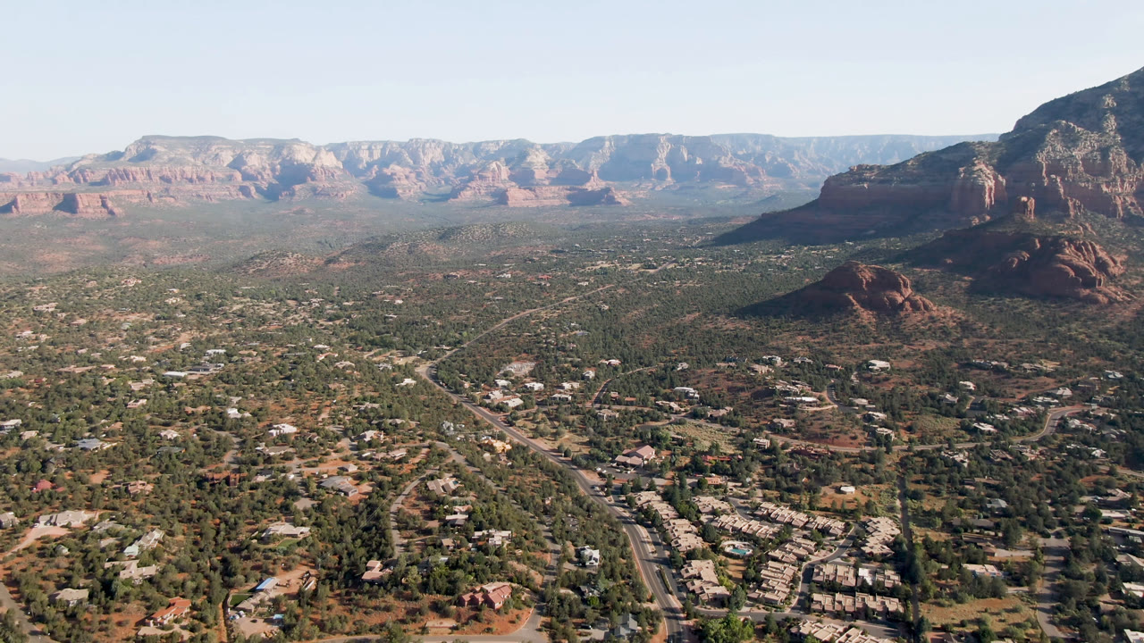 Aerial frame over Sedona, Arizona, with a large red rock butte near the right side, green valley terrain below, visible roads, and no visible people.
