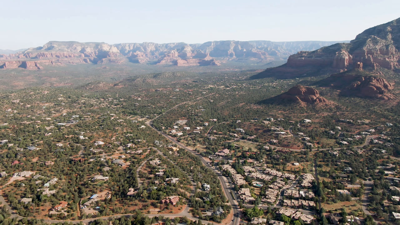 High aerial view of Sedona, Arizona, showing low buildings and roads in the foreground, green desert valley in the middle distance, red rock cliffs, and no visible people.