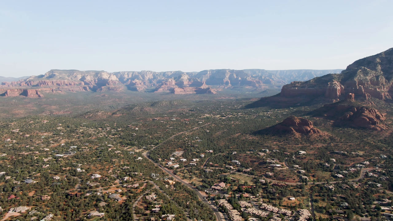 Second-frame aerial over Sedona, Arizona with a diagonal road and low buildings below, red cliffs stretching across the horizon, a right-side rock mass, and no visible people.