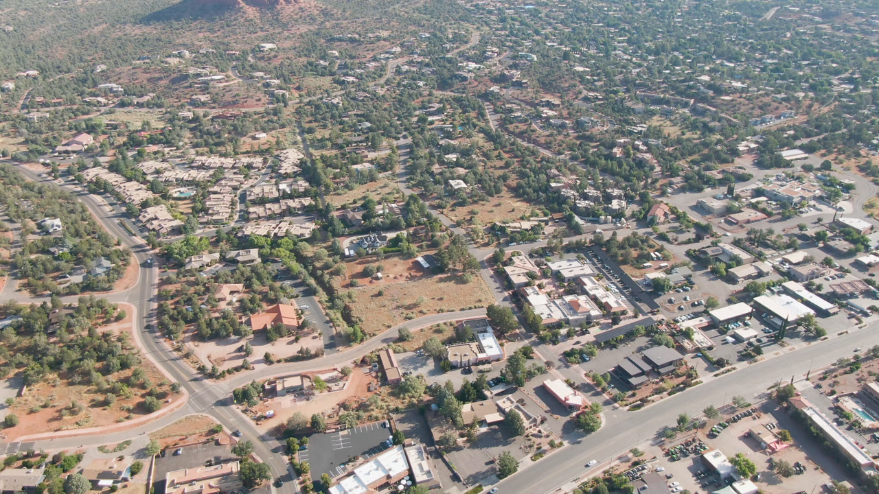 Opening aerial frame over Sedona, Arizona with curving roads, low buildings, parked cars, green trees, reddish ground, hazy hills, and no visible people.