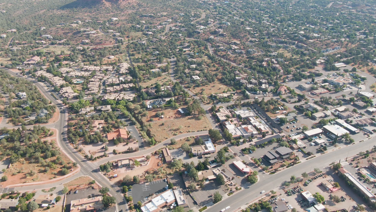 Second aerial frame over Sedona, Arizona showing a slightly shifted view of rooftops, parking areas, curved streets, dense trees, dry lots, and no visible people.