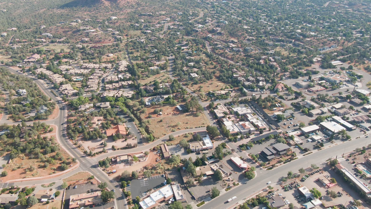 Mid-clip aerial frame over Sedona, Arizona with a broad road across the lower frame, small commercial buildings, tree-lined neighborhoods, dry open ground, and no visible people.