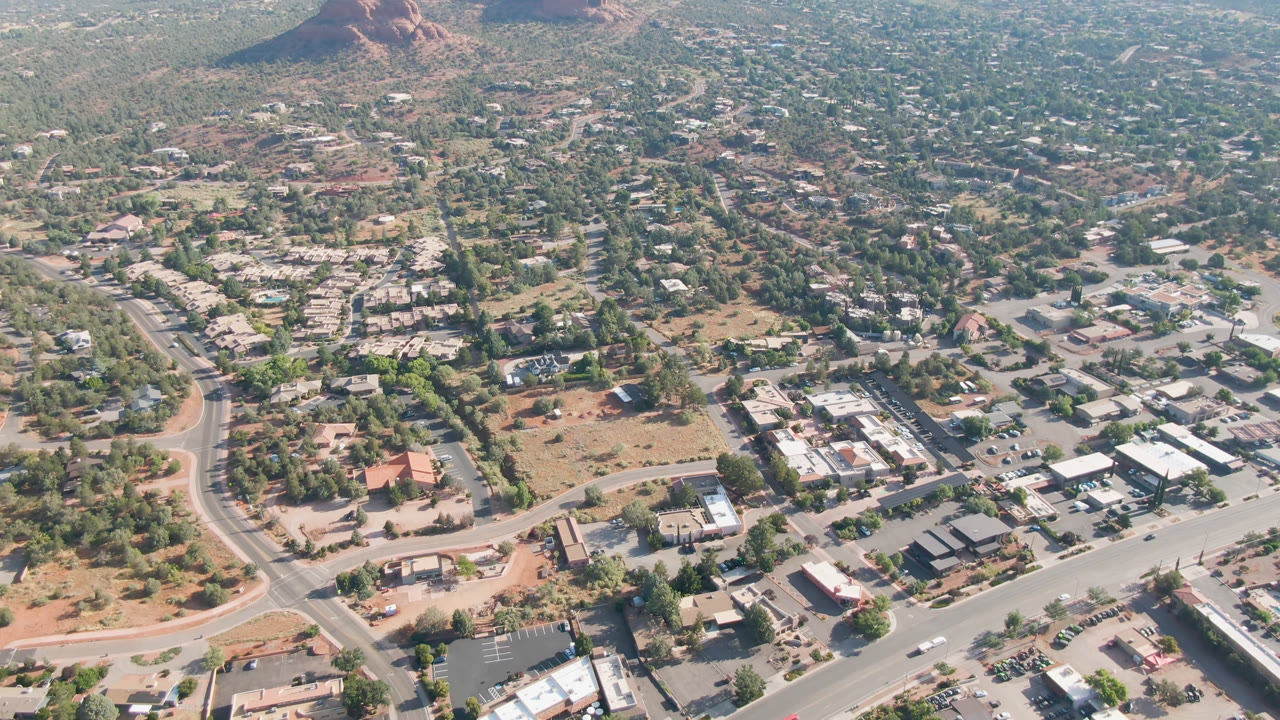 Opening aerial frame over Sedona, Arizona, with a curved road, scattered low buildings, desert trees, and red rock hills in the distance, with no visible people.