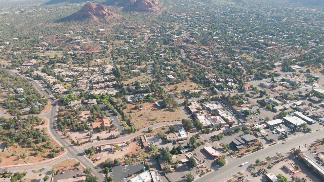 One second into the clip, the aerial view holds over Sedona streets, small commercial-looking buildings, tree cover, dry lots, and red rock formations beyond town, with no visible people.