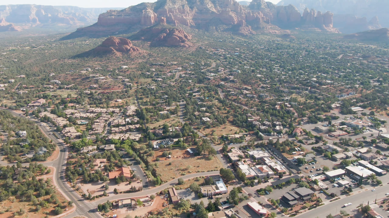 Opening aerial frame over Sedona, Arizona, with roads and low buildings below, desert vegetation across town, red rock formations in the distance, and no visible people.