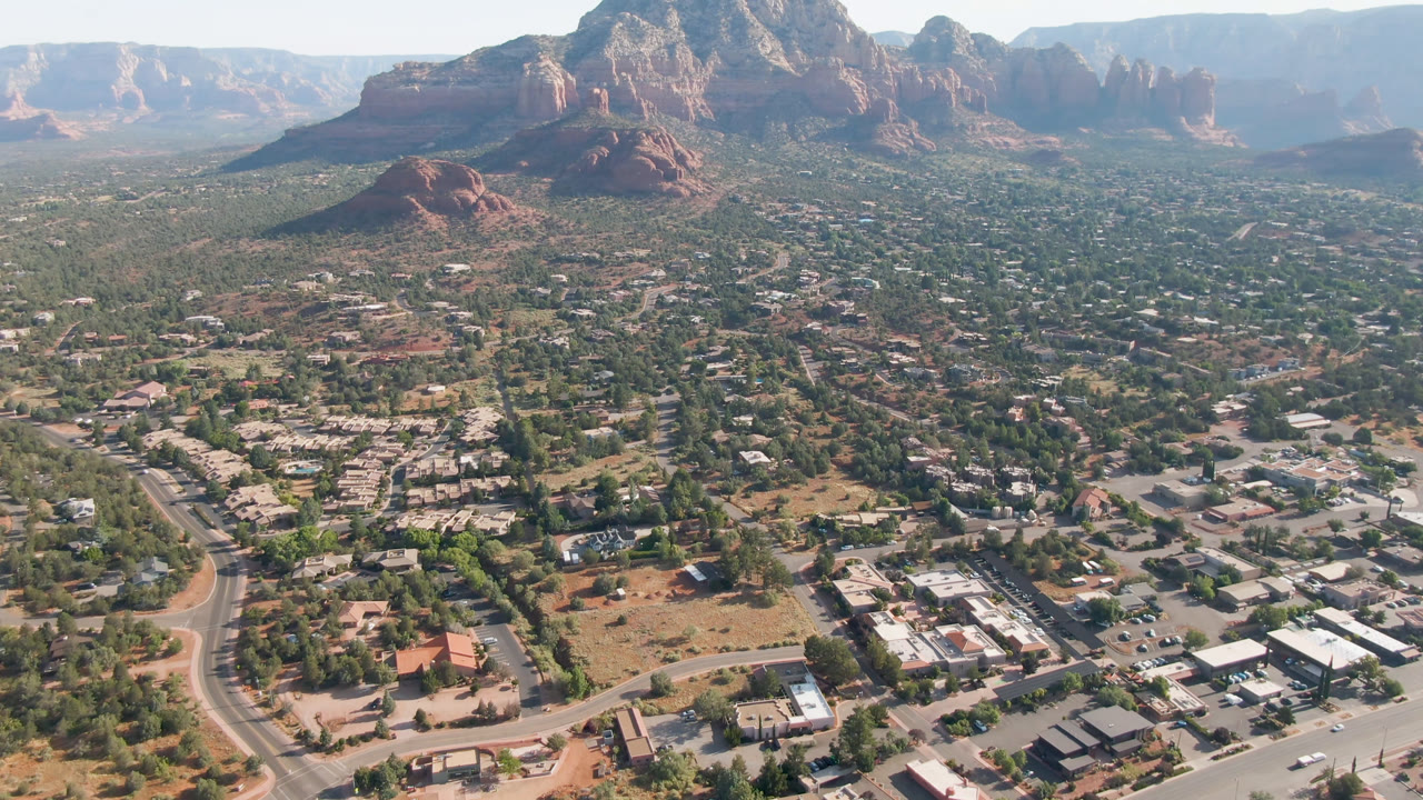 Aerial frame over Sedona, Arizona, showing a winding road at lower left, clusters of buildings, green desert scrub, red rock buttes, and no visible people.