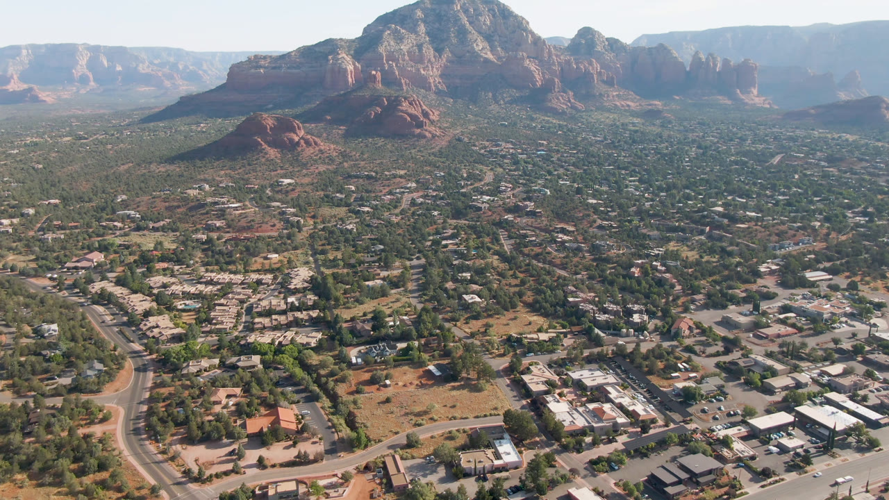 Mid-clip aerial frame over Sedona, Arizona, with town blocks and roads in the lower frame, scattered vegetation through the middle, layered red rock formations behind, and no visible people.