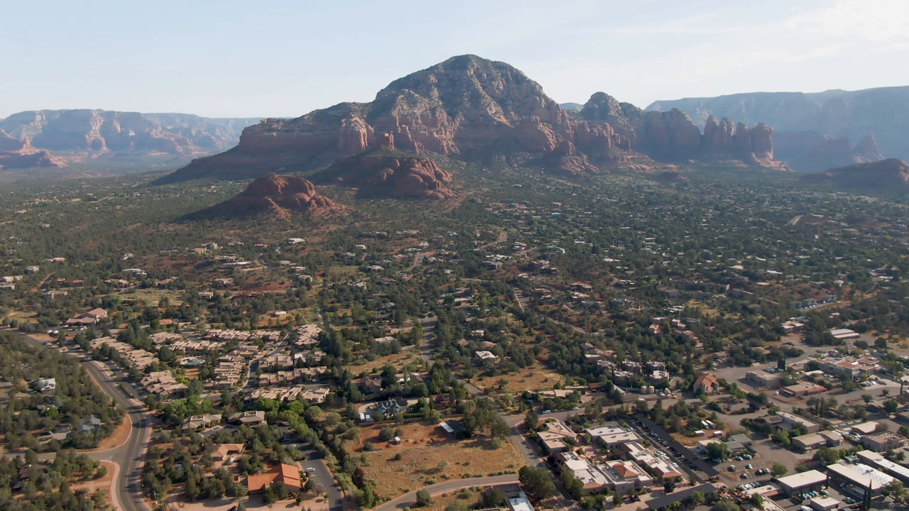 Opening frame over Sedona, Arizona with roads and low buildings in the foreground, red rock bluffs in the distance, pale sky above, and no visible people.