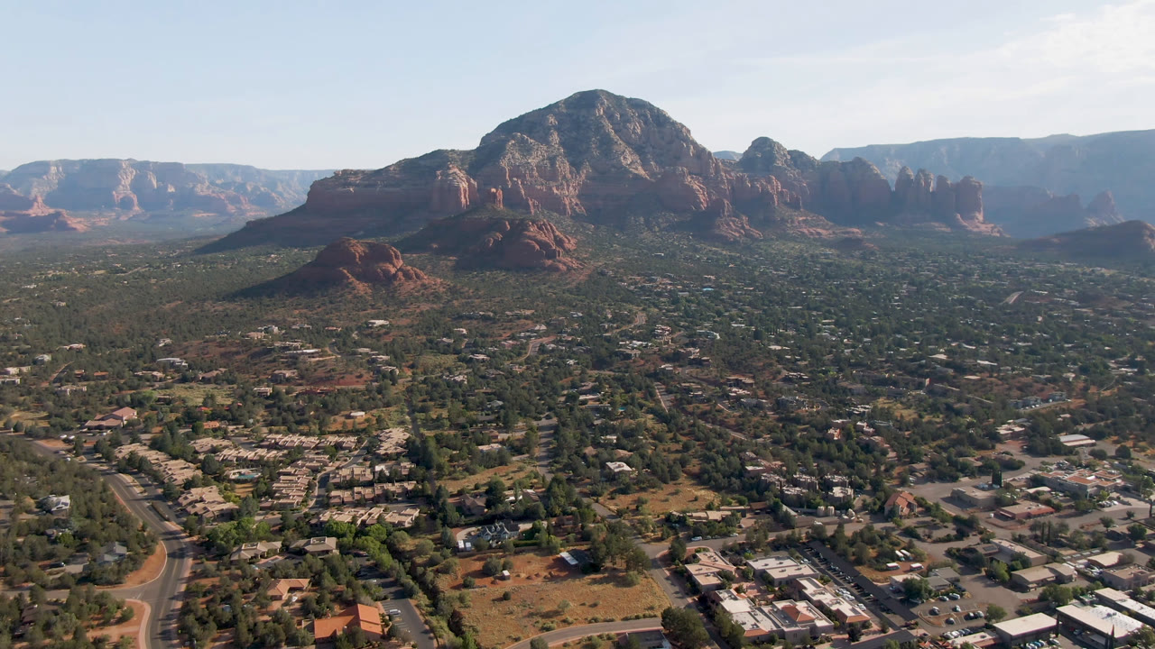 Second frame from the Sedona aerial holds a central red rock mass above scattered buildings, road lines, scrubland, distant ridges, and no visible people.