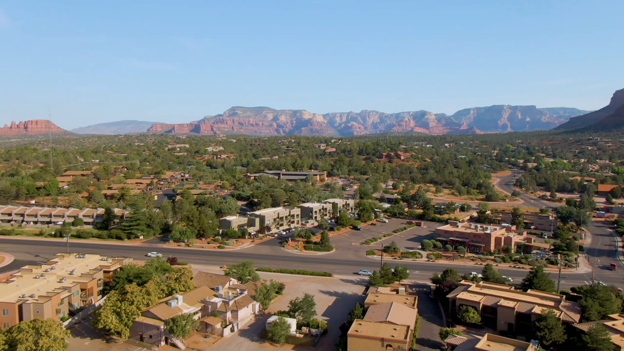 Opening aerial frame over Sedona, Arizona, showing tan rooftops, intersecting roads, scattered trees, distant red rock formations, and no visible people.