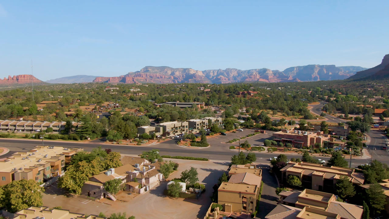 Mid-clip aerial frame of Sedona, Arizona, with a multi-lane road, small vehicles, low commercial and residential buildings, tree cover, red rock cliffs, and no visible people.