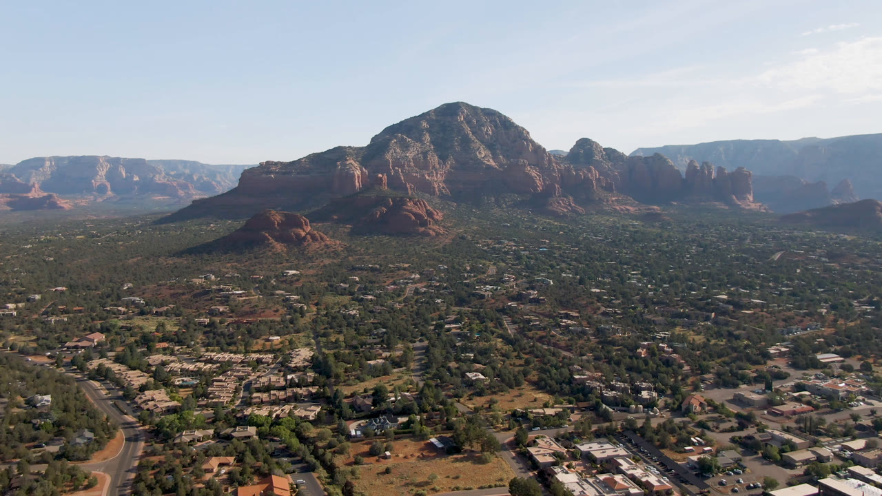 Opening frame over Sedona, Arizona, with roads, trees, low buildings, a large red rock bluff, distant ridges, pale sky, and no visible people.