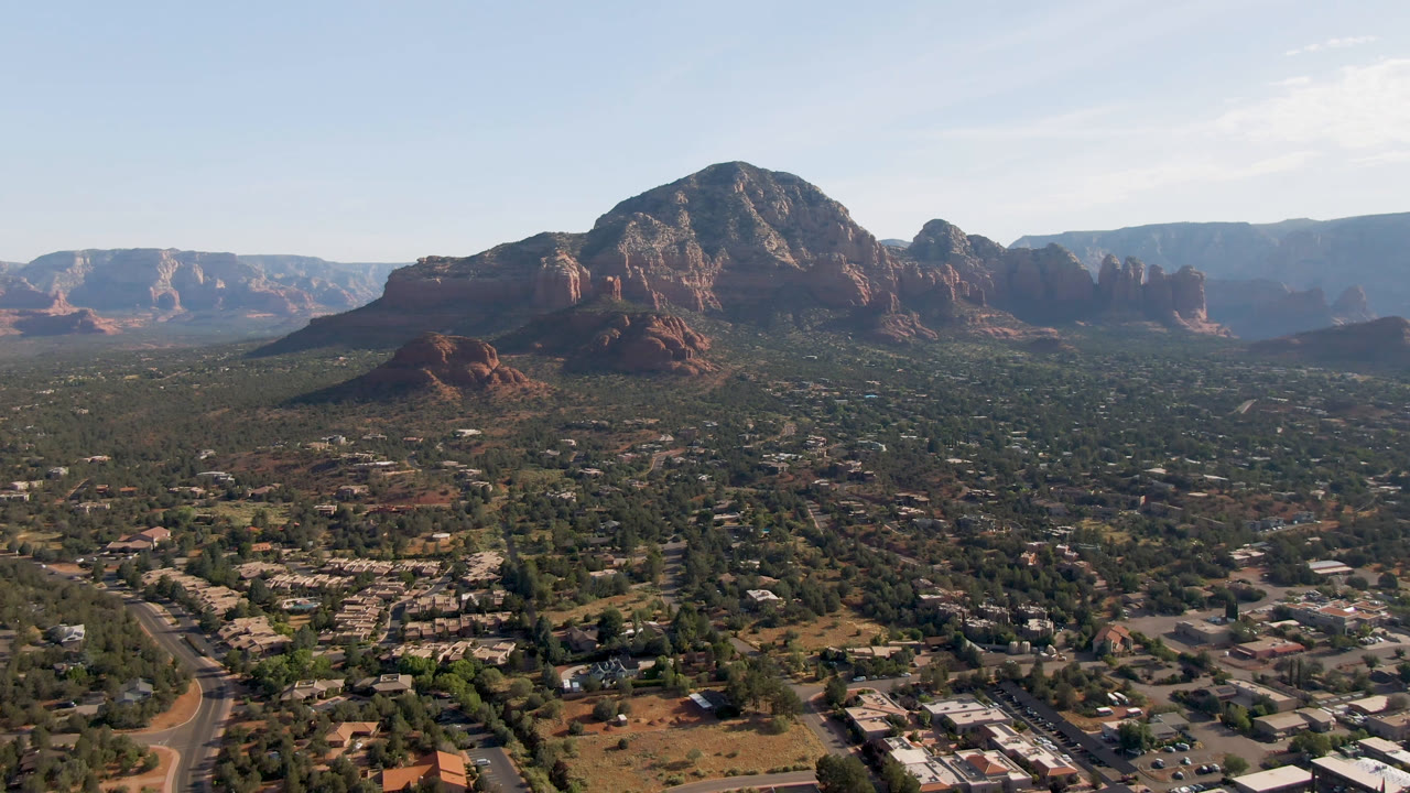 Second frame of the Sedona aerial holds a broad view of residential streets, desert vegetation, red rock formations, distant mountains, soft daylight, and no visible people.