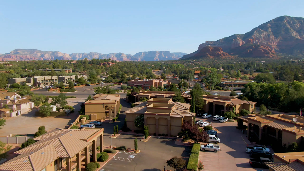 Opening aerial frame of Sedona, Arizona showing tan buildings, a parking lot with scattered cars, green trees, red rock mountains, blue sky, and no visible people.