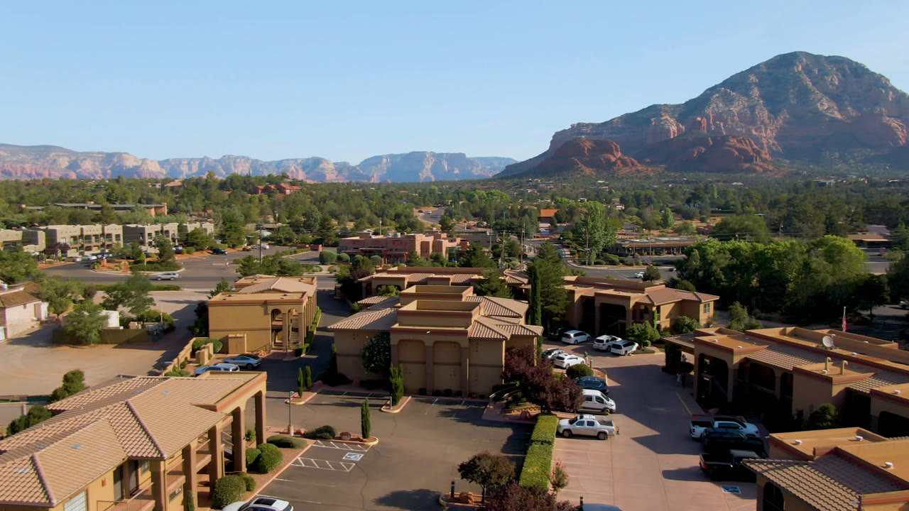 Second aerial frame of Sedona, Arizona with tiled rooftops and parking spaces in front, tree-lined streets in the middle, red rock mountains behind, and no visible people.