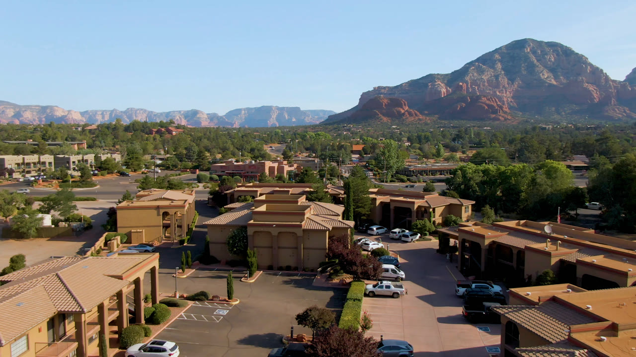 Third frame of the Sedona, Arizona aerial view showing a broad parking lot, tan rooflines, scattered vehicles, green trees, red rock formations, and no visible people.
