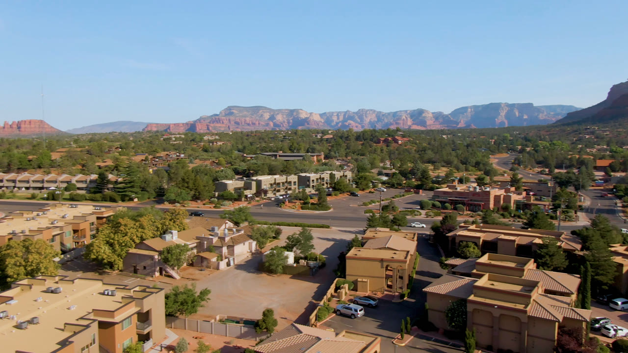 Opening aerial frame over Sedona, Arizona with tan rooftops and streets in the foreground, green trees across town, red rock ridges on the horizon, and no visible people.