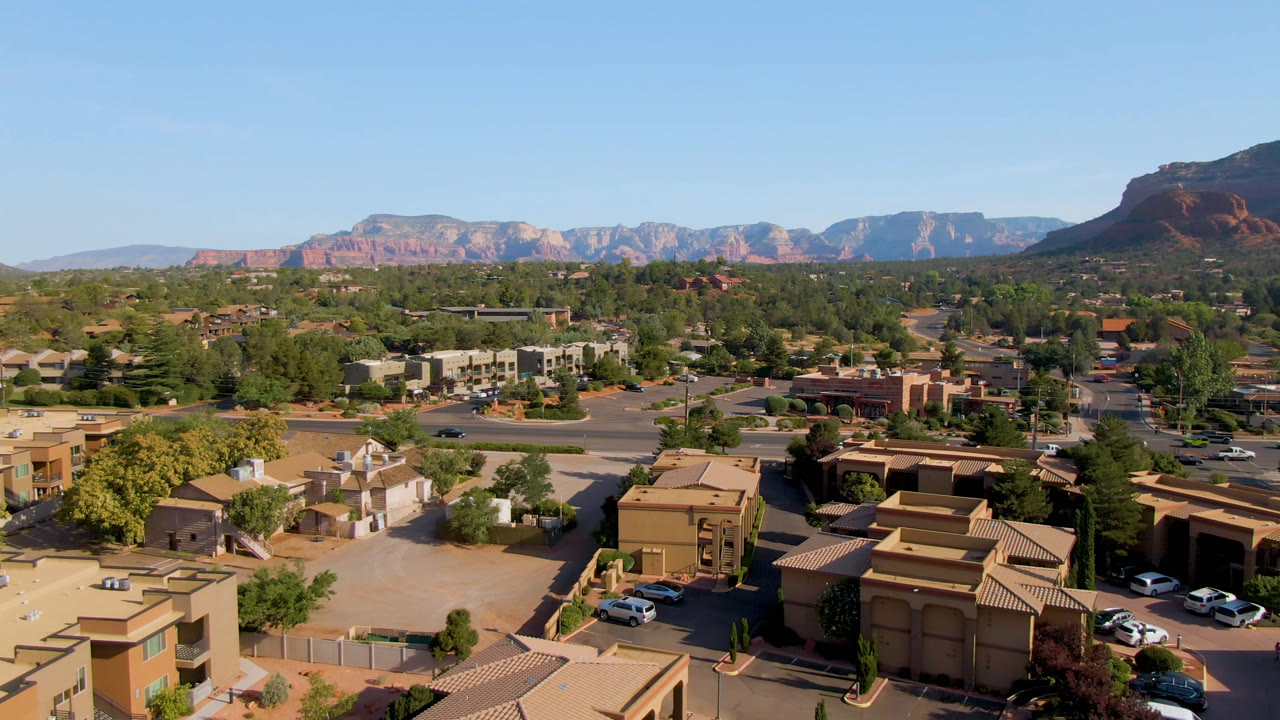 Second aerial frame of Sedona, Arizona showing low buildings and parking areas below, tree-lined streets through the center, distant red rock cliffs, and no visible people.