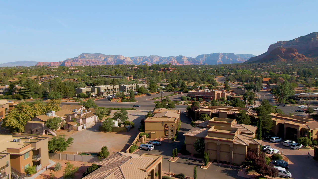 Third aerial frame over Sedona, Arizona with sunlit tan buildings, small parking lots, a broad road, dense trees, a dark mountain shoulder to the right, and no visible people.