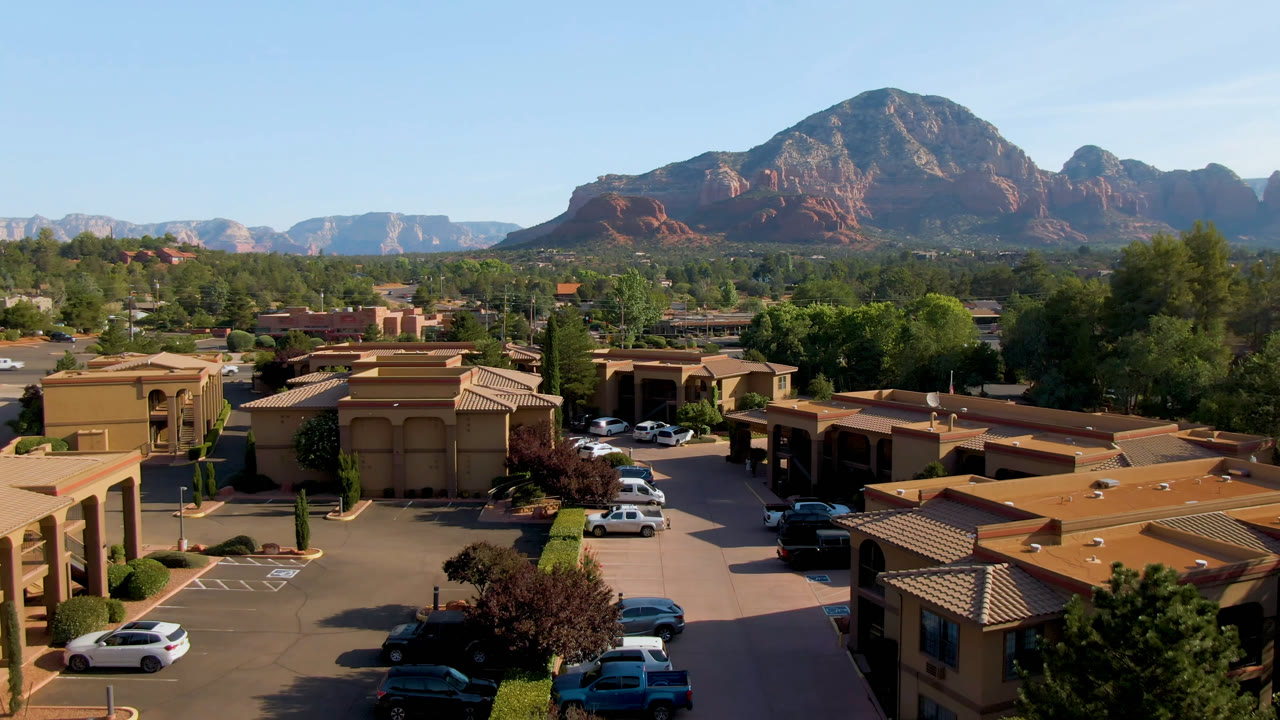 Opening frame of Sedona, Arizona from above, with tan buildings, parked cars, trees, red rock mountains in the distance, clear sky, and no visible people.