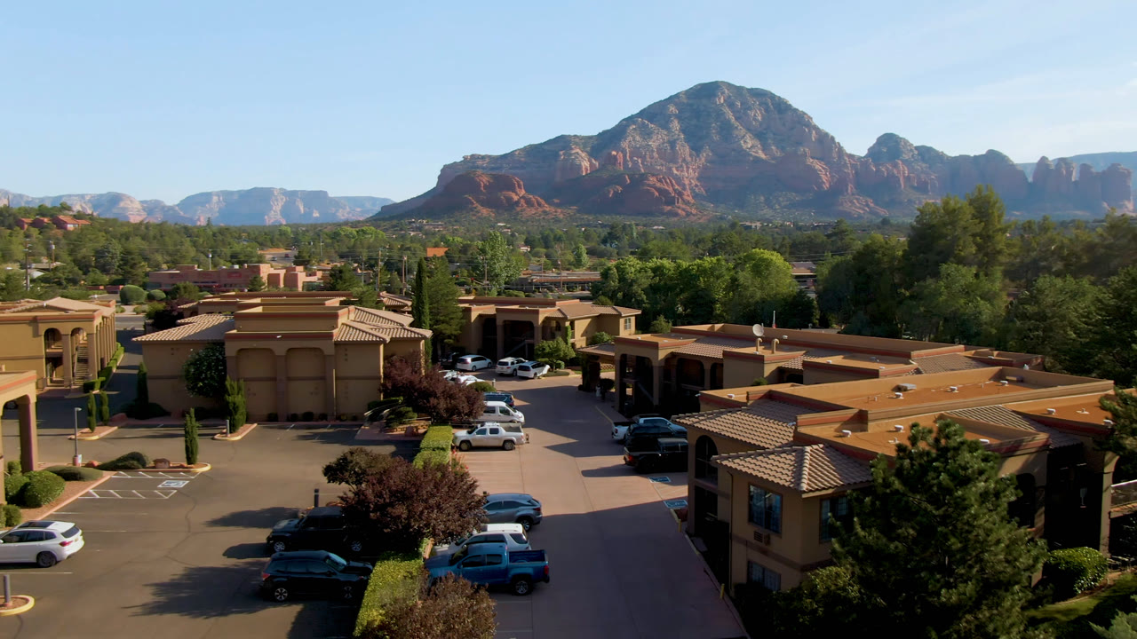 Third frame from the Sedona, Arizona aerial view, with foreground rooftops, a small parking lot, dense trees, distant red mountains, clear daylight, and no visible people.