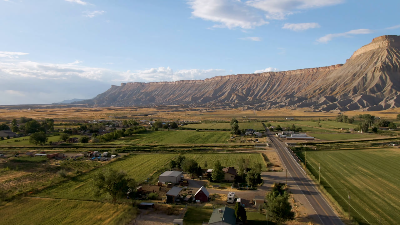 Opening frame over rural Palisades, Colorado, showing green fields, a straight road, scattered farm structures, and a long mesa ridge with no visible people.