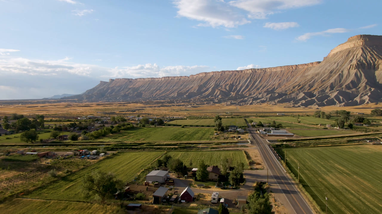 Second frame of rural Palisades, Colorado, with rectangular fields, roadside buildings, tree clusters, and the mesa ridge stretched across the background with no visible people.