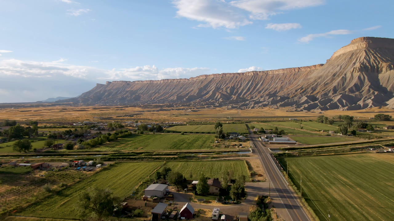 Aerial landscape with road, green fields, scattered buildings, and mesa cliffs.