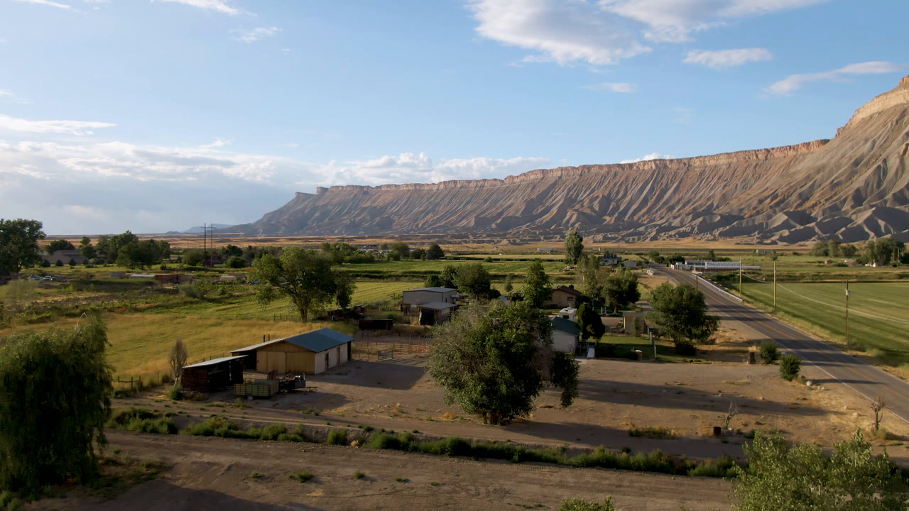 Elevated rural valley view with fields, homes, road, trees, and mesa ridge.