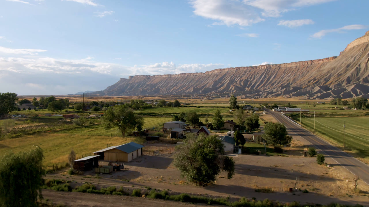 Second frame of Palisades, Colorado with a straighter view down the road, rural buildings in the foreground, farmland in the midground, mesa ridges behind, and no visible people.