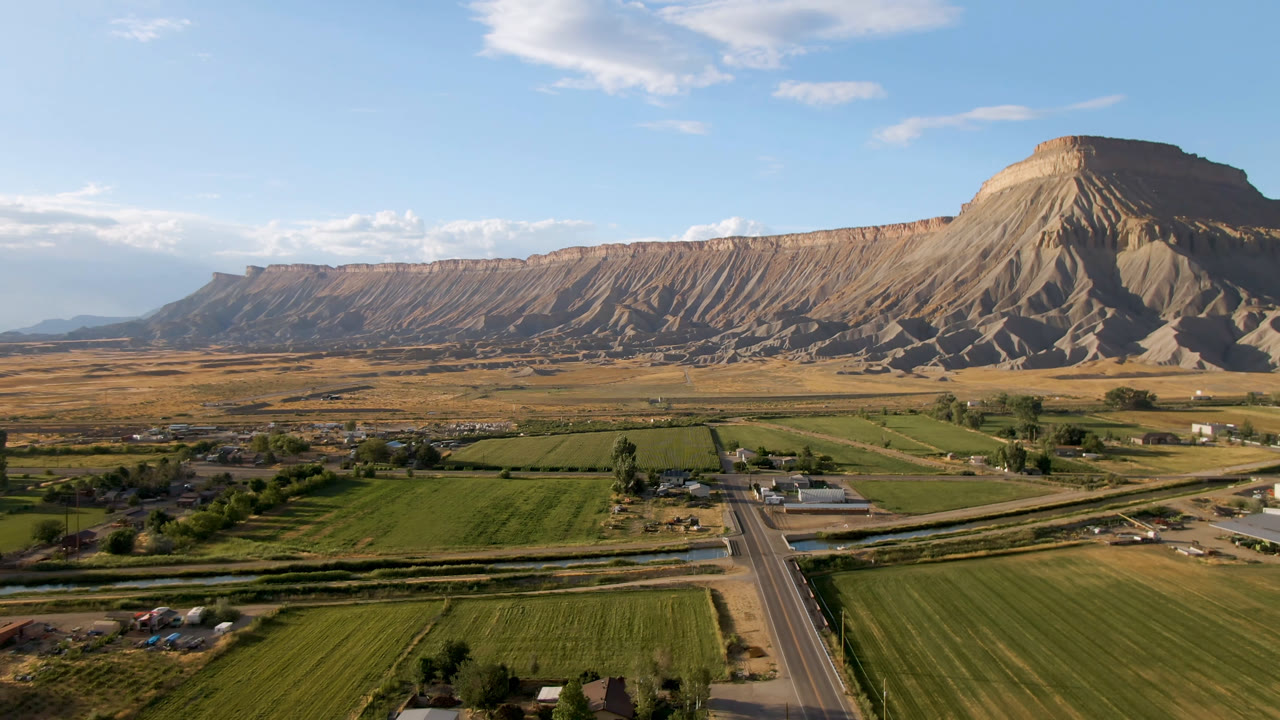 Wide elevated view in Palisades, Colorado, with green fields and a road in the foreground, scattered buildings below a large mesa, and no visible people.
